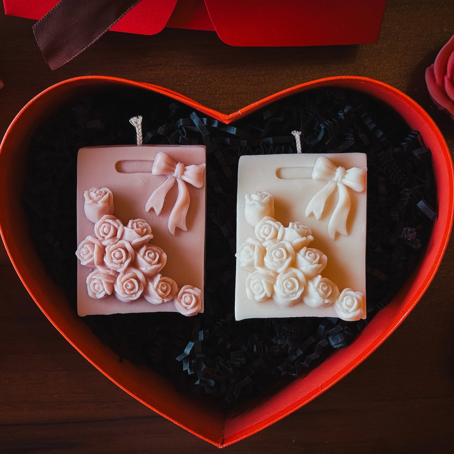 Two decorative candles shaped like a gift bag with decorative flowers and bows in a heart-shaped red box.