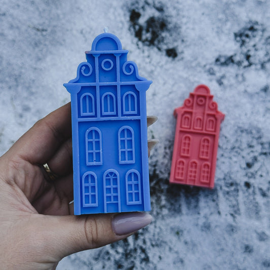 Blue and red Amsterdam house-shaped candles held by a hand on a textured surface.