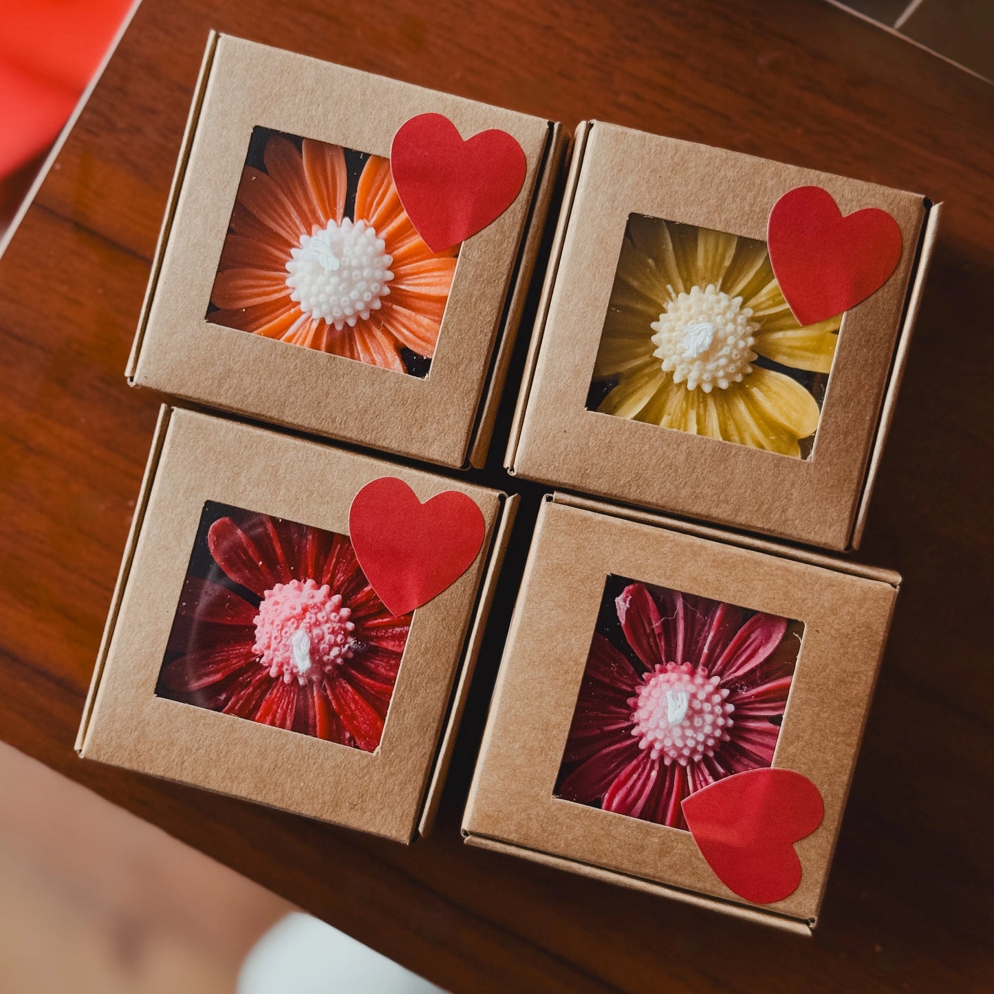 Four brown gift boxes with windowed panels packing daisy candles with a red heart sticker, displayed on a wooden surface.