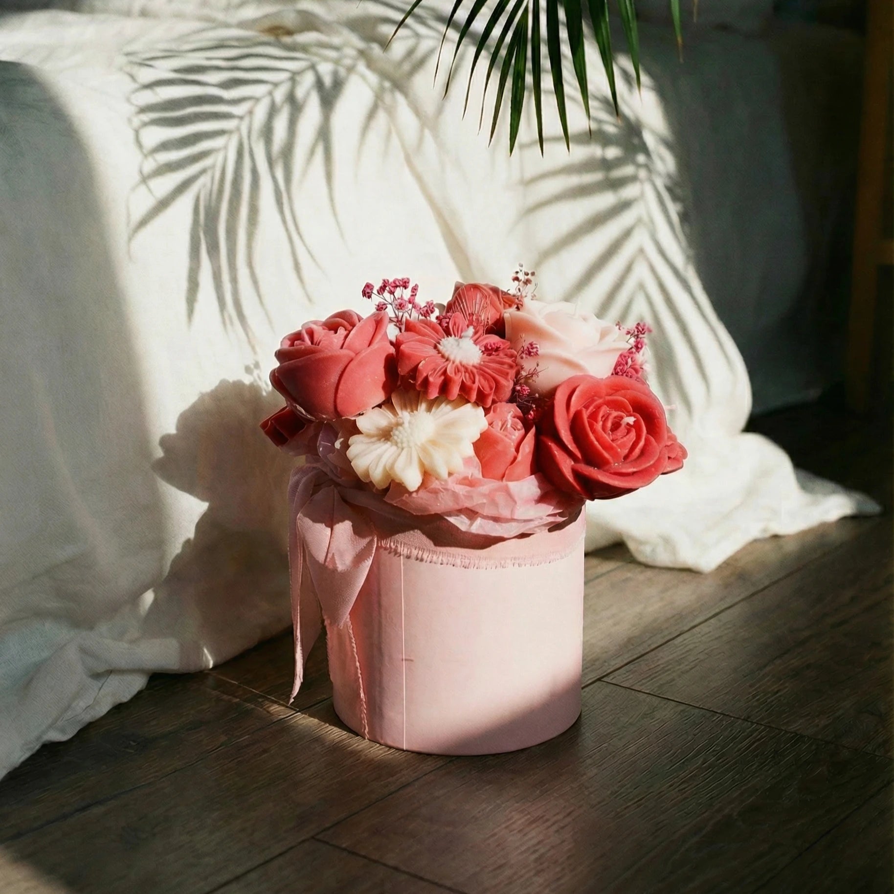 Pink candle flower arrangement in a decorative pink pot on a wooden floor with white cushions and a plant in the background.
