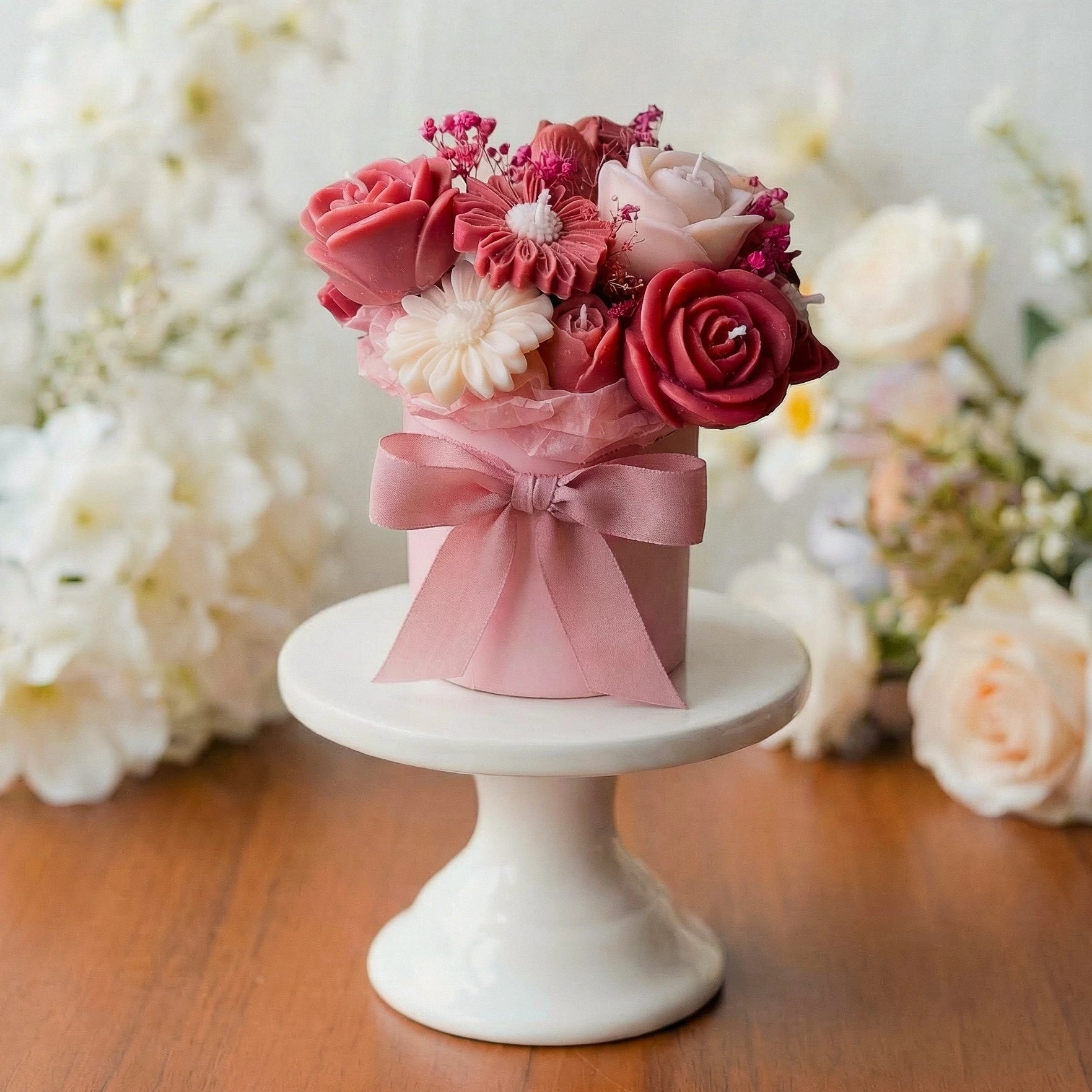 Bouquet of pink and red wax flowers with a pink ribbon on a white pedestal against a floral background.