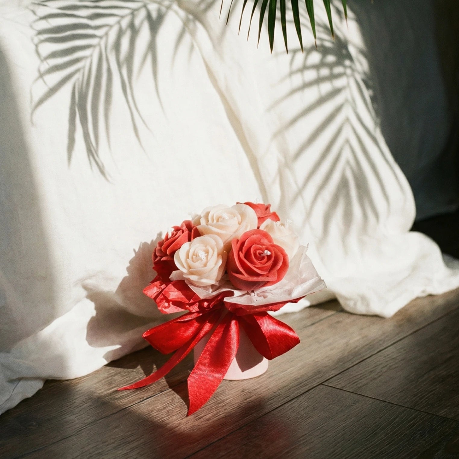Bouquet of red and white wax flowers with a red ribbon on a wooden surface, with white fabric and green leaves in the background.