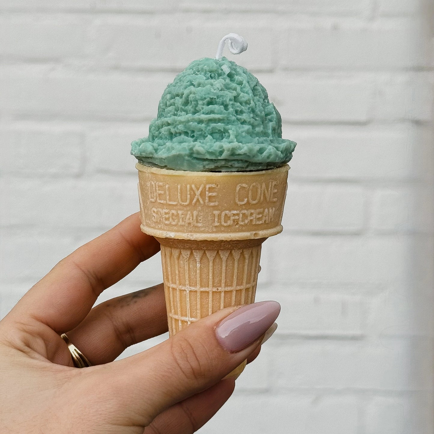 Candle shaped like a green ice cream cone in a waffle cone held by a hand against a light gray brick wall.