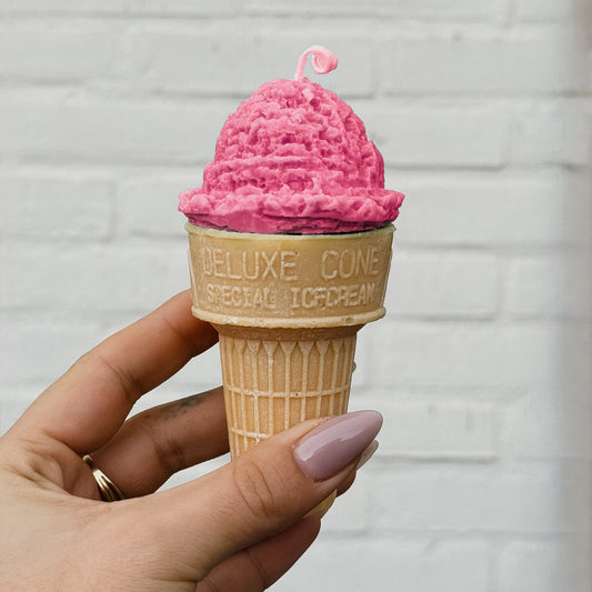 Candle shaped like a pink ice cream cone in a waffle cone held by a hand against a light gray brick wall.