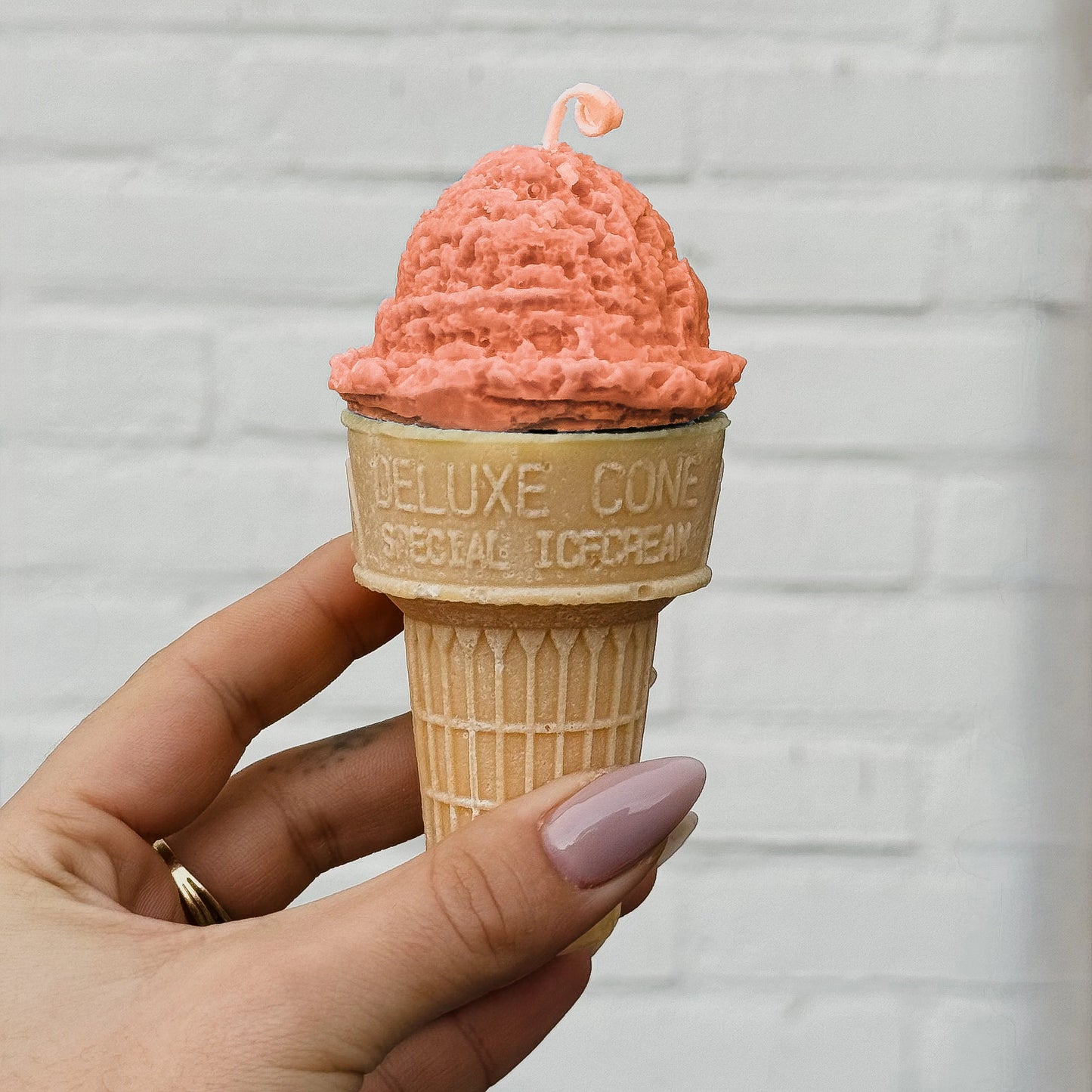 Candle shaped like an orange ice cream cone in a waffle cone held by a hand against a light gray brick wall.